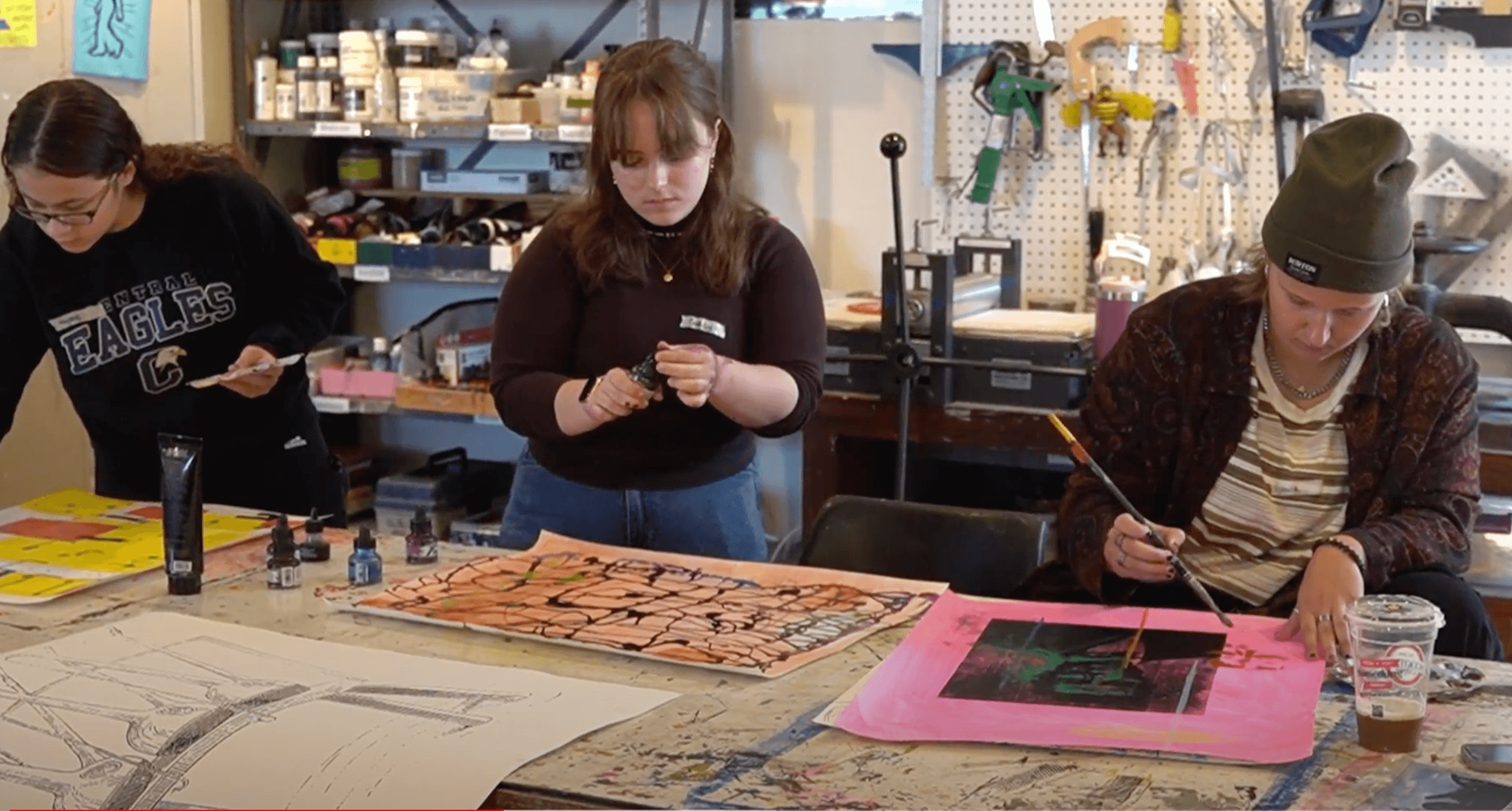 Three people in an art studio are working on their projects. The studio is filled with supplies. One person uses ink, another holds a tool, and the third paints. Various artworks are laid out on the table.