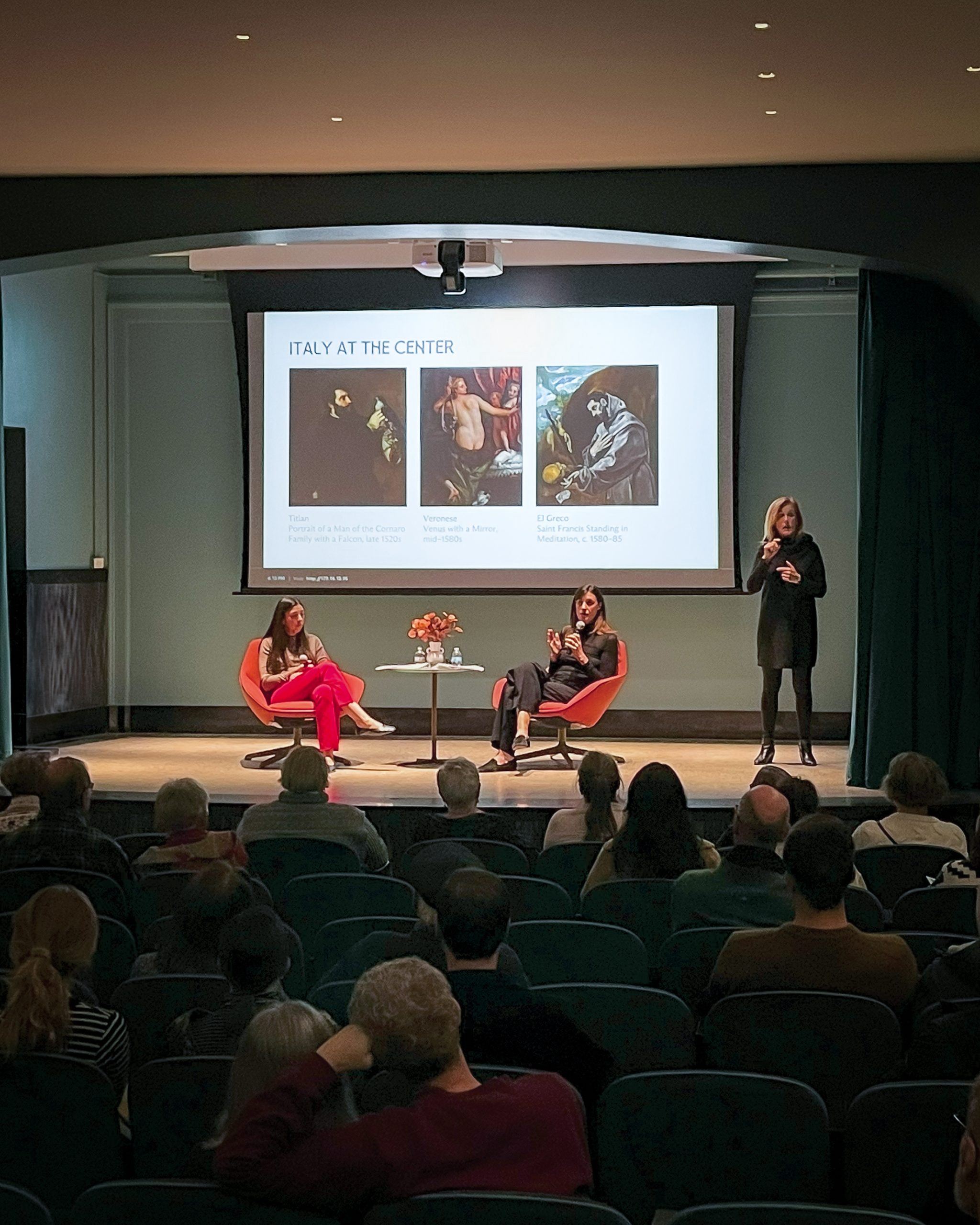 A speaker on stage delivers a presentation titled "Italy at the Center" to an audience. Two seated panelists are onstage near a screen displaying images and text. The audience watches attentively in a dimly lit auditorium.
