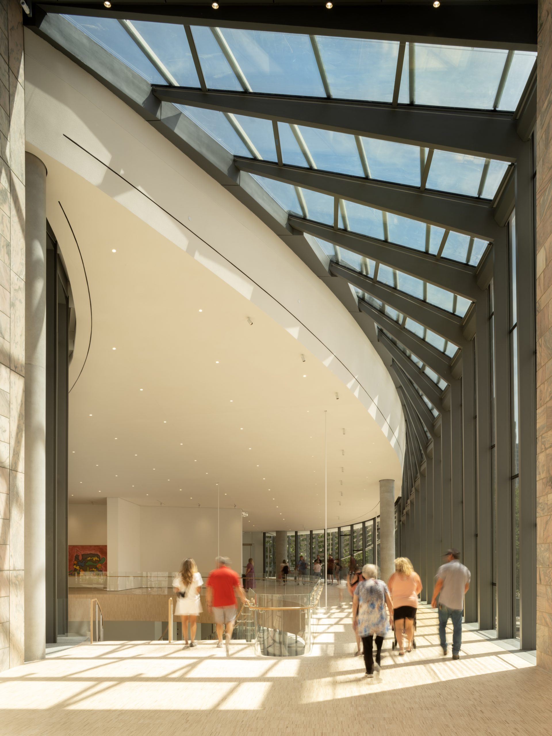 Interior of a spacious modern building with a high, curved ceiling and large glass windows. People are walking along a path. Natural light floods the space through the skylights above.