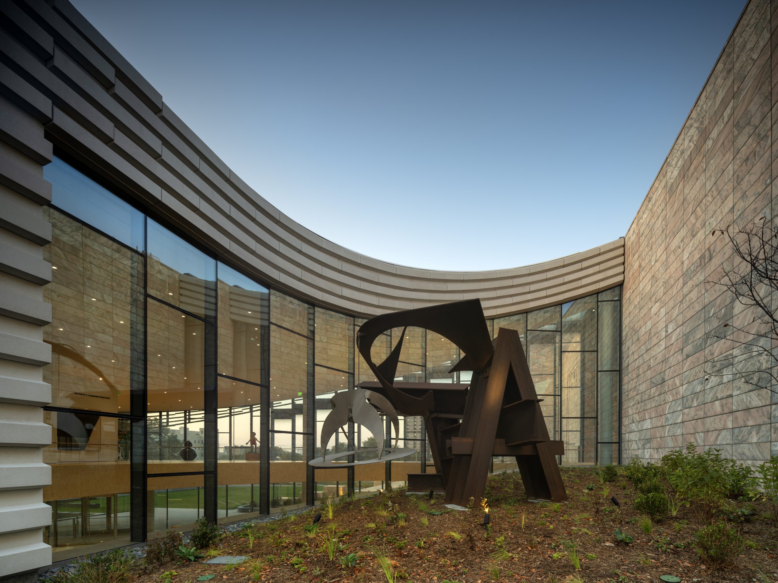 A large abstract metal sculpture stands in a landscaped courtyard outside a modern building with tall glass windows and textured stone walls under a clear sky.