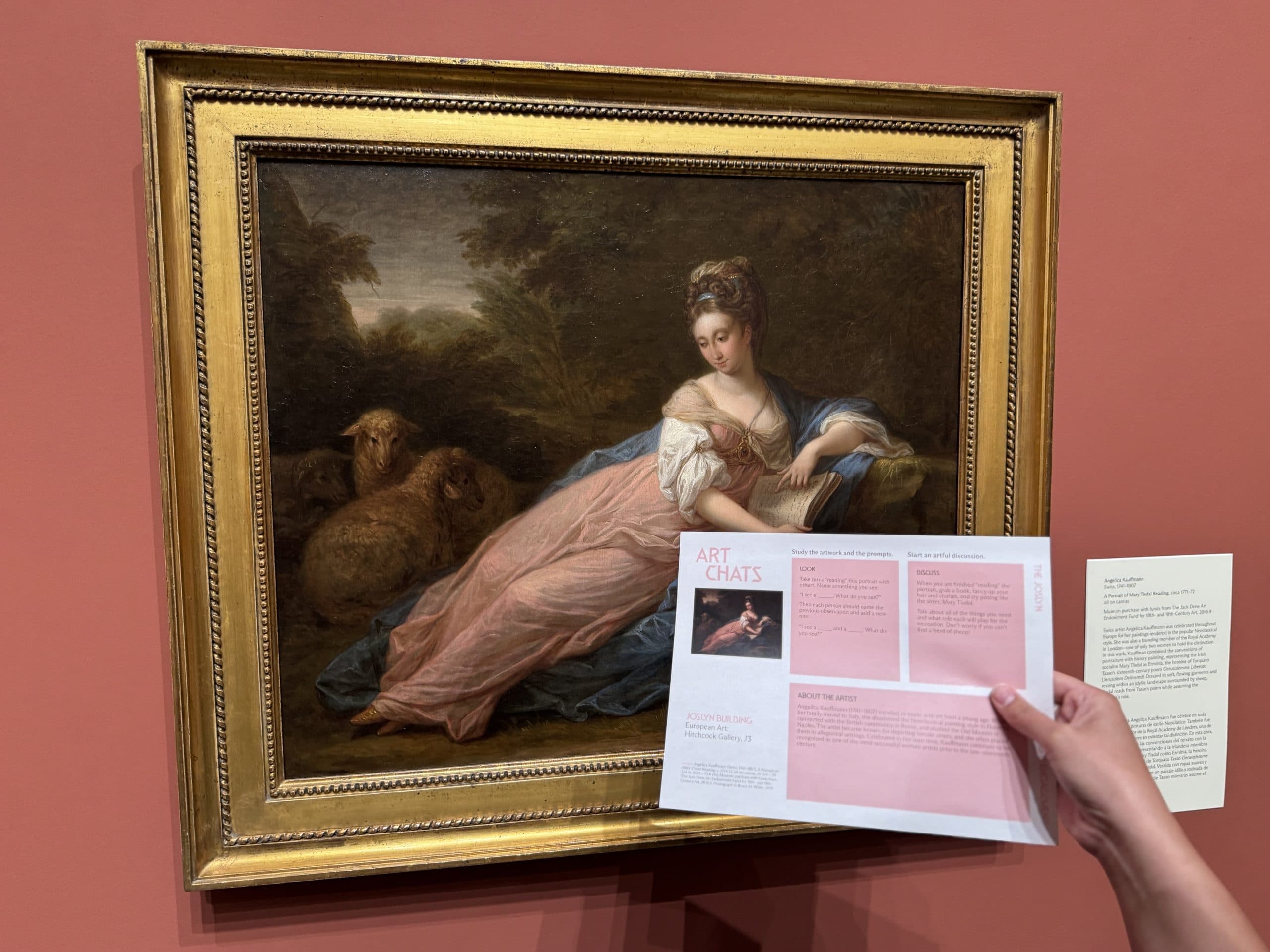 A person holds a pink and white museum brochure in front of a framed painting of a woman in 18th-century clothing, sitting outdoors with sheep, displayed on a red wall with an info card beside it.