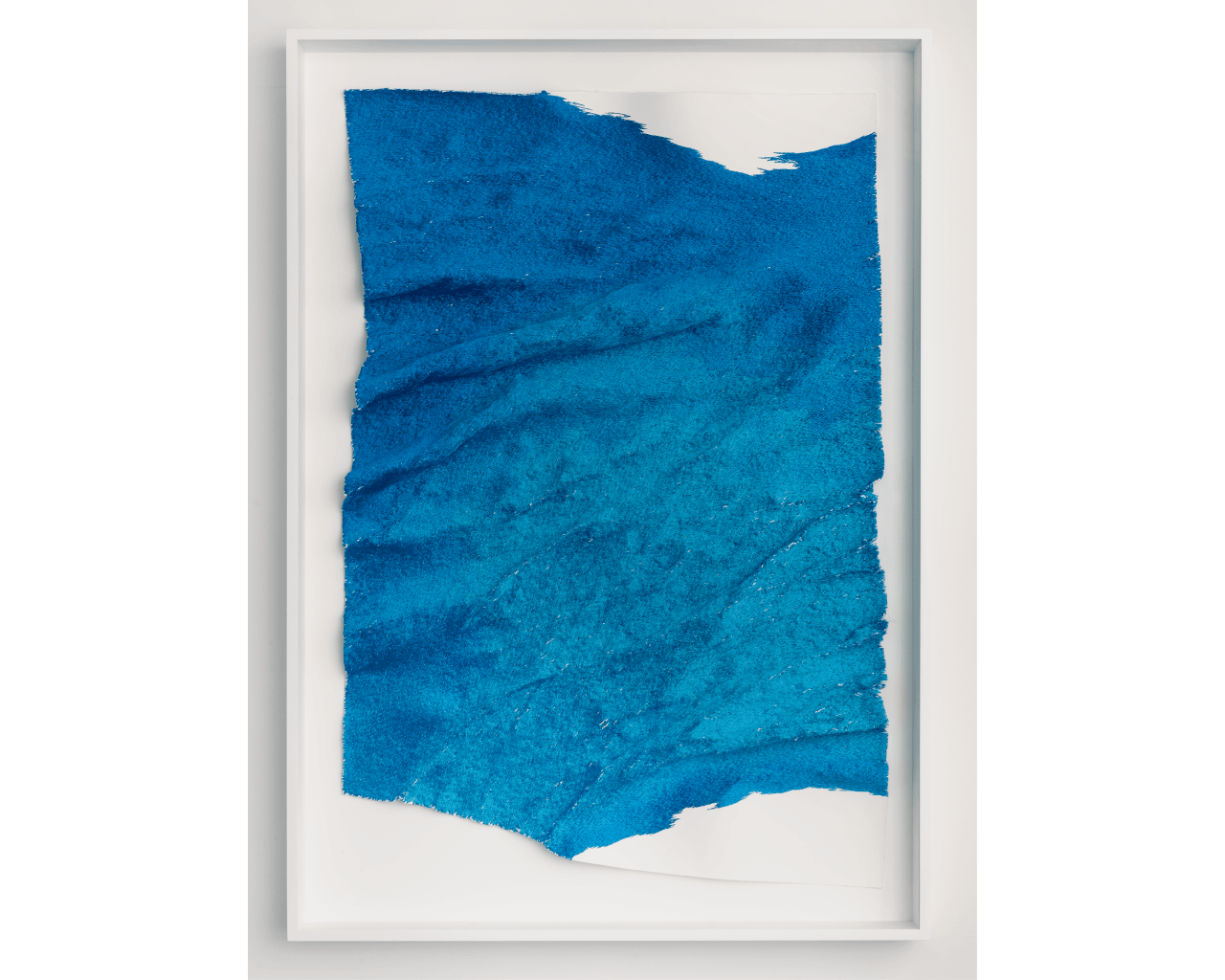 A textured, torn sheet of vibrant blue material is framed under glass with a white mat and frame. The uneven edges and varied blue tones create an abstract, wave-like appearance.