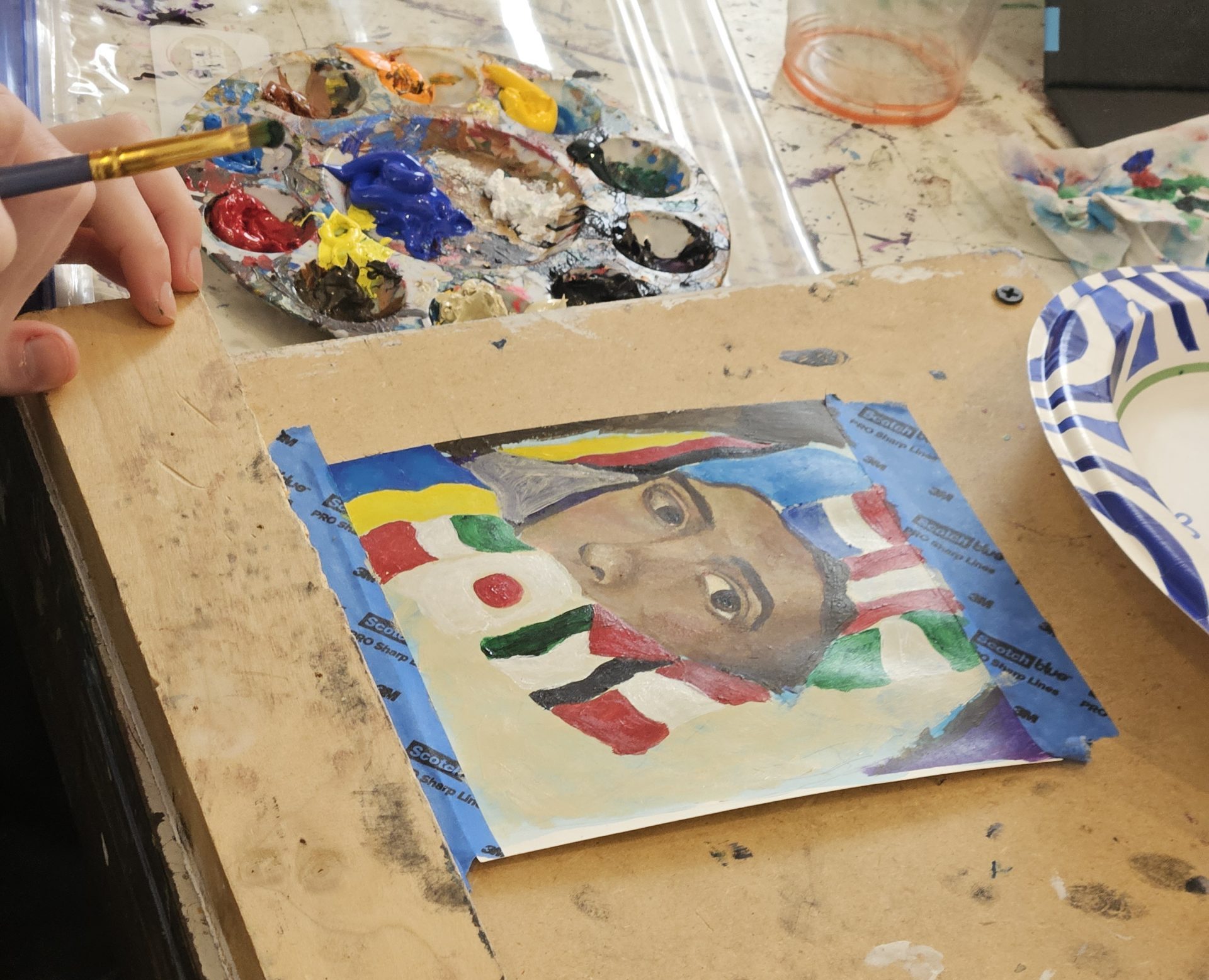 A person paints a colorful portrait of a face on paper, surrounded by paint palettes and brushes. A smartphone on the table displays various country flags, and a paper plate sits nearby.