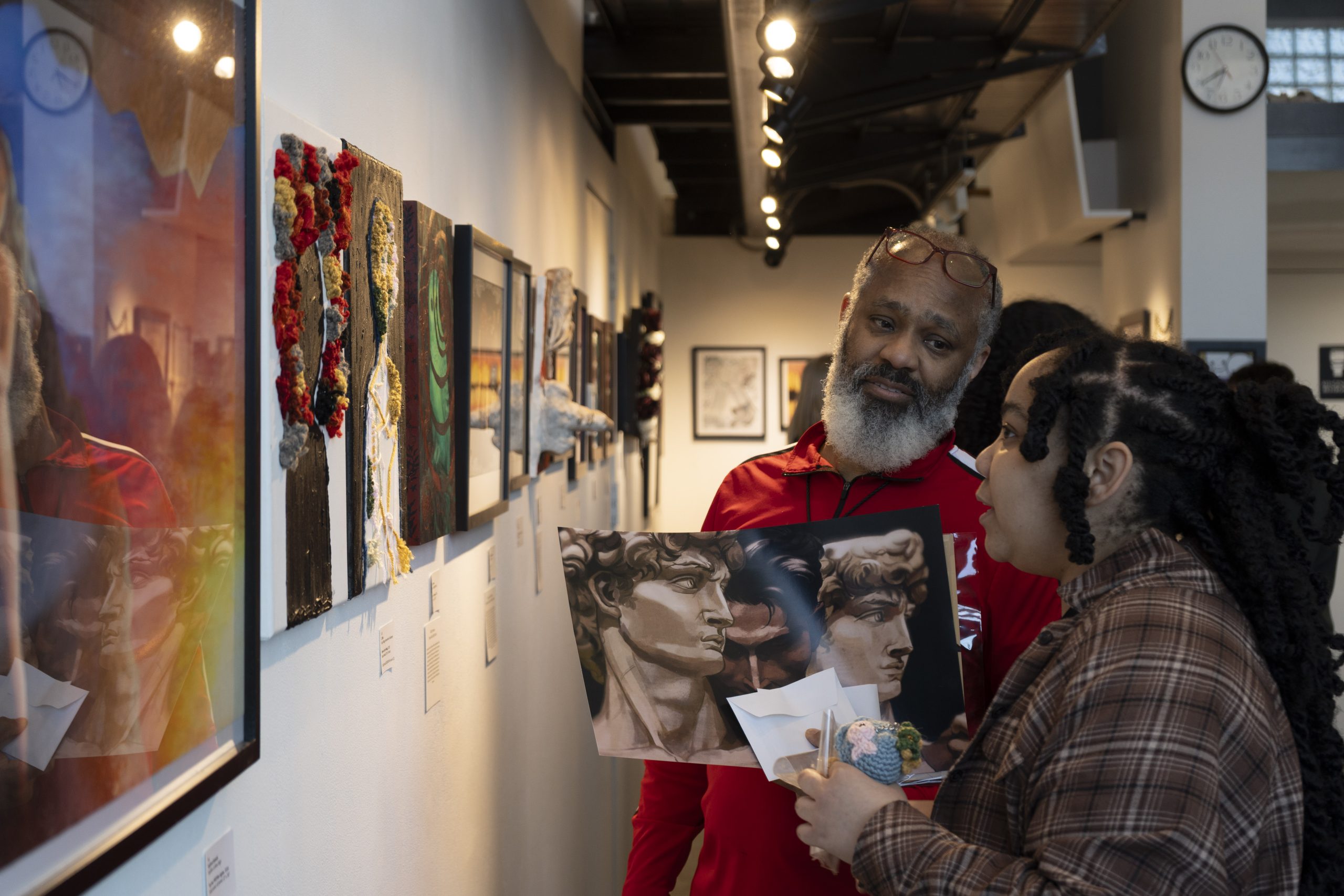 A man and a woman stand in an art gallery, looking at paintings on the wall. The woman holds a collage artwork and appears to be discussing it with the man, who listens attentively. Bright lights illuminate the gallery.