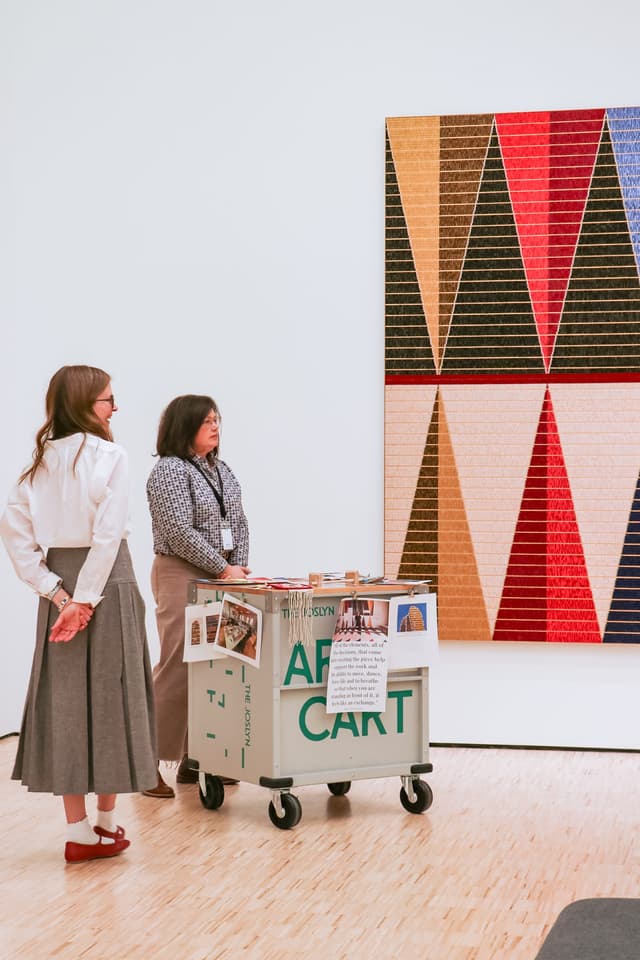 Two women stand near an art cart in a gallery, looking at a large geometric artwork with red, black, beige, and gold triangles on the wall. The cart holds art materials and displays informational signs.