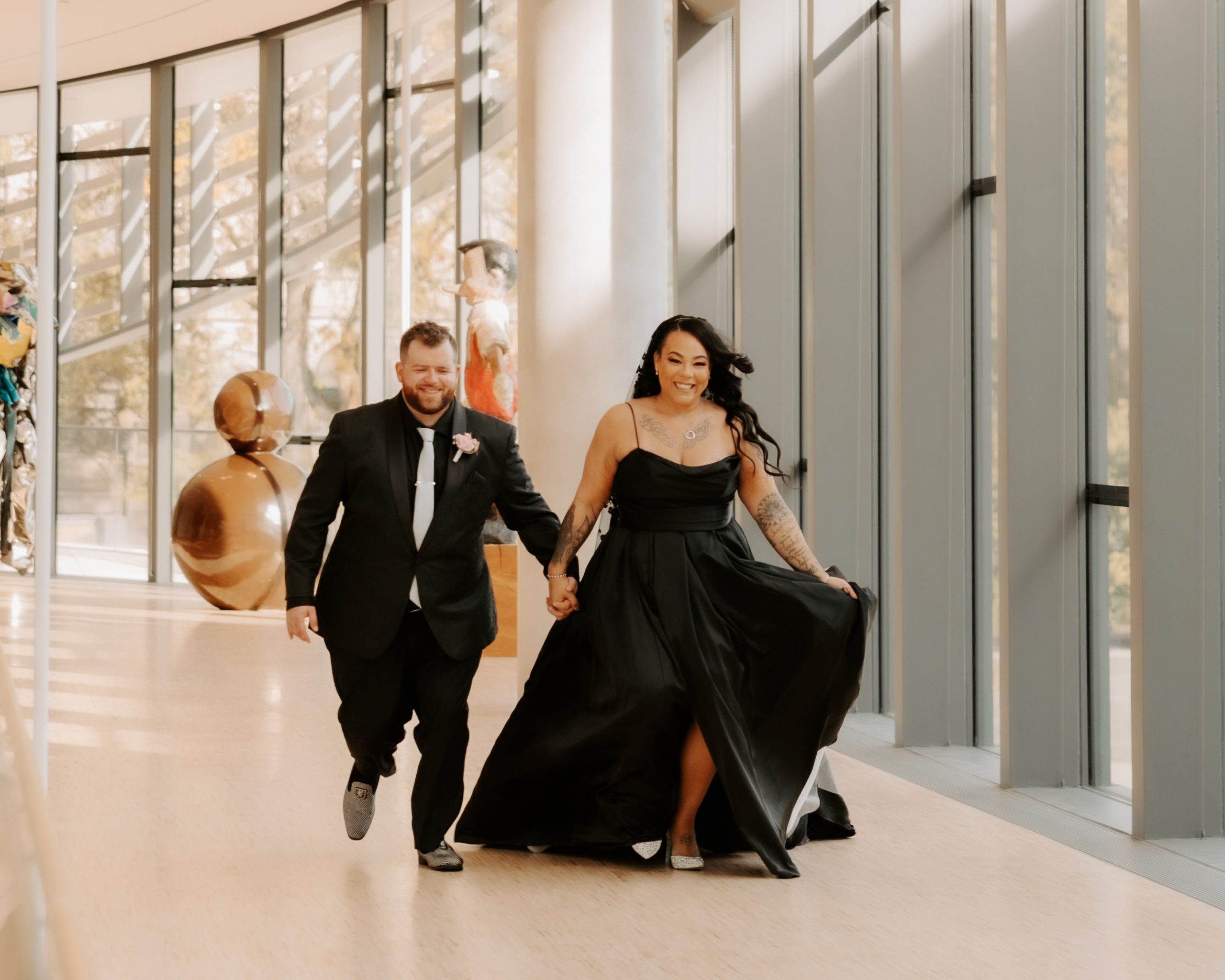A couple is joyfully walking hand in hand down a sunlit glass corridor. The woman wears a flowing black dress awend the man is in a black suit with a tie. Both are smiling as they move towards the camera.