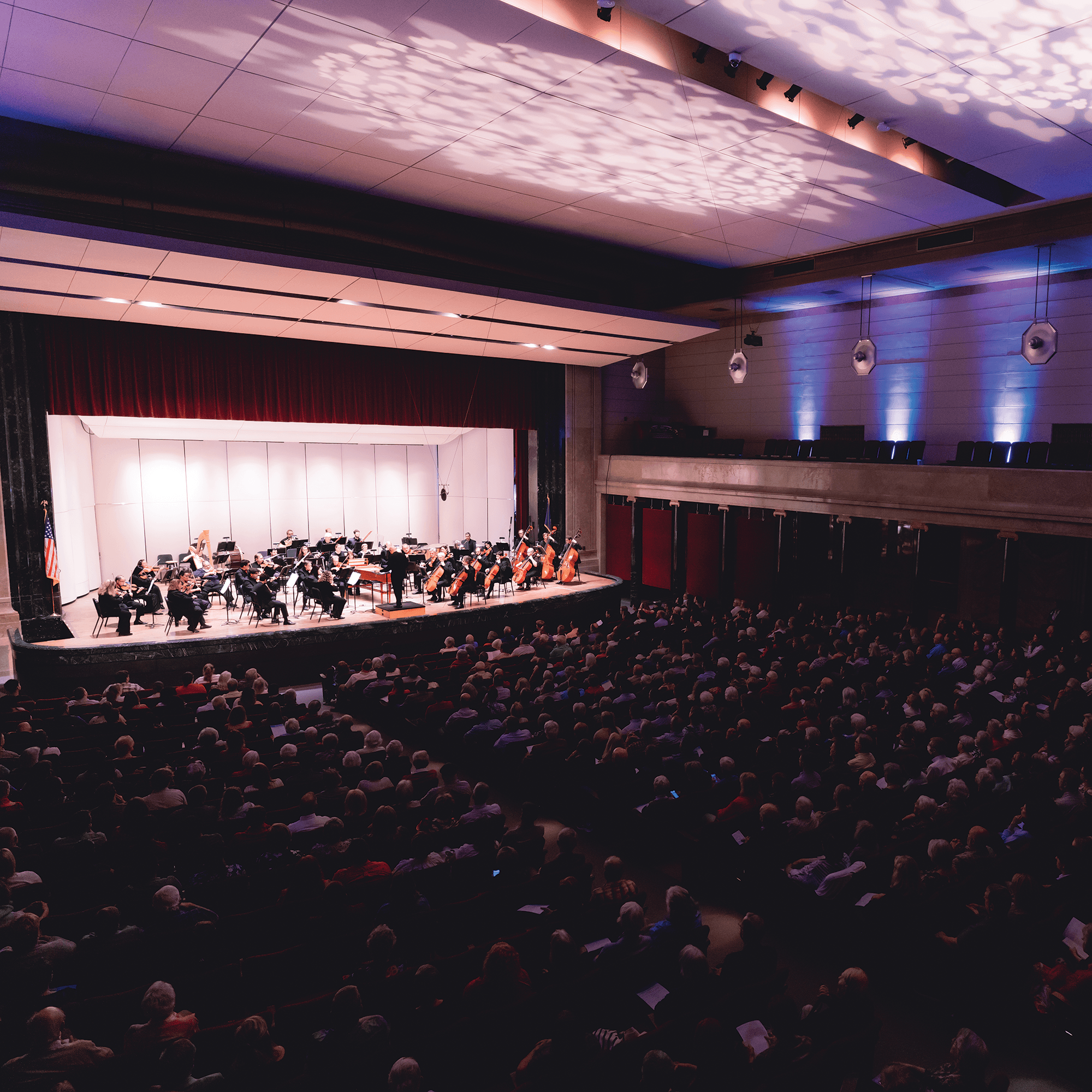 An orchestra performing on a brightly lit stage in a concert hall. The audience is seated in rows, watching the performance. The stage is framed by tall red curtains, and the ceiling has decorative lighting patterns.