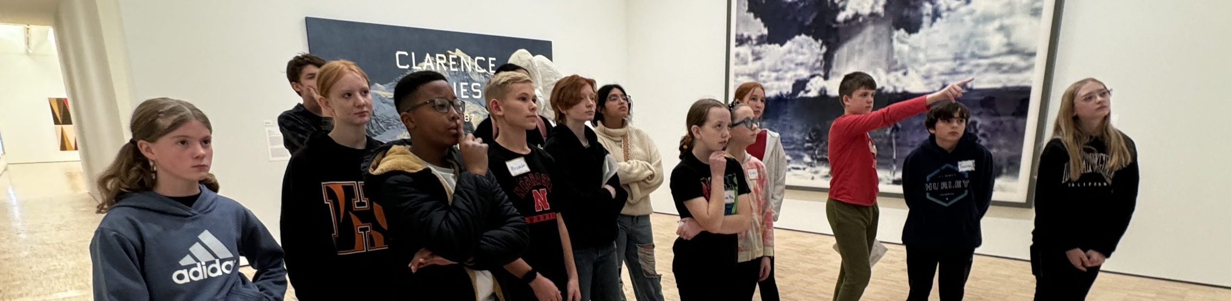 A group of teenagers stands together in an art gallery, attentively looking at artwork on the wall ahead. One person is pointing, and the setting has bright lighting with modern wooden floors.
