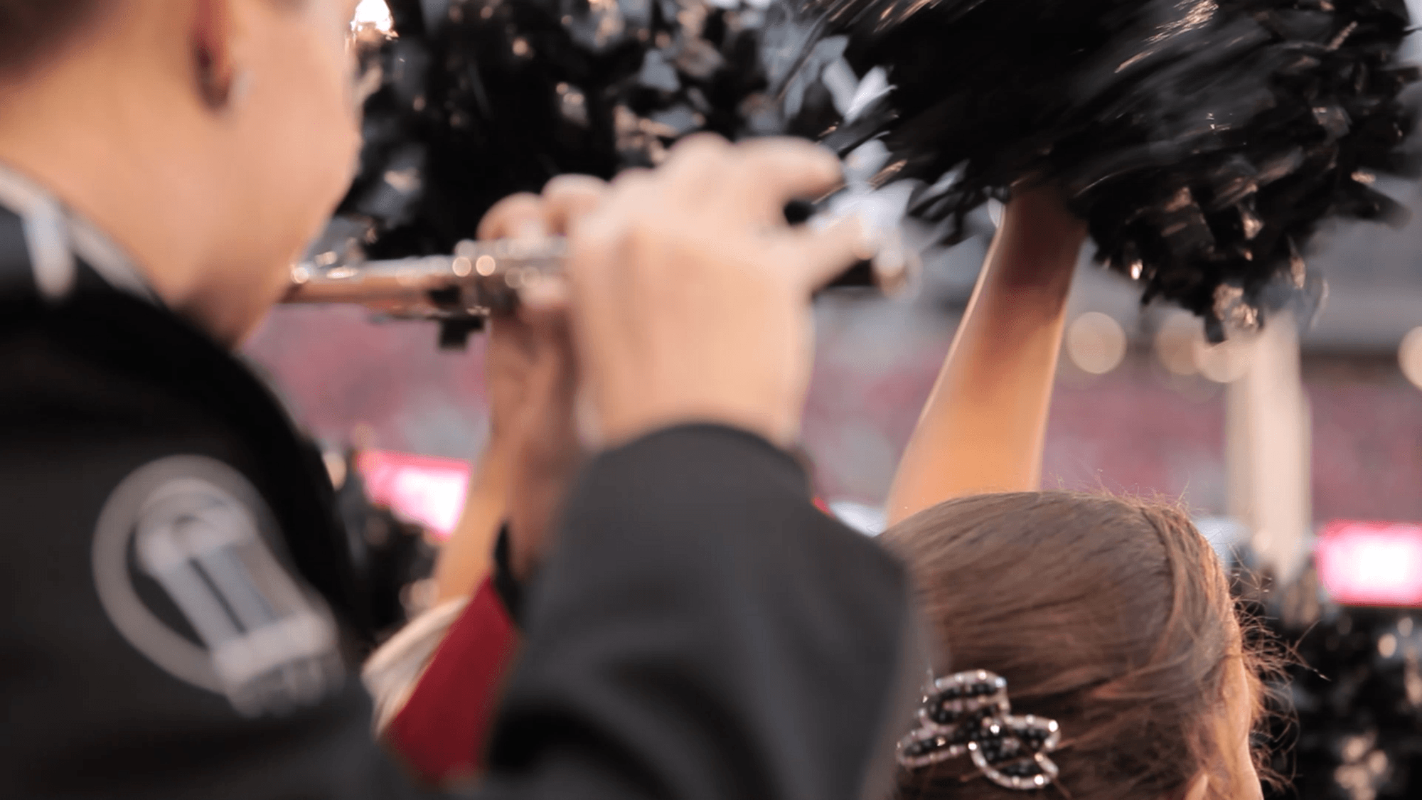 A close-up of a marching band member playing a flute next to a cheerleader holding black pom-poms, with a blurred crowd and stadium lights in the background.