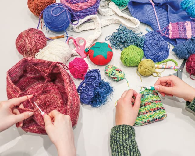 Two people crochet with colorful yarn. Various yarn balls, crocheted pieces, scissors, and a strawberry-shaped pincushion are scattered on a table. One person works on a pink project, another on a multicolored piece.