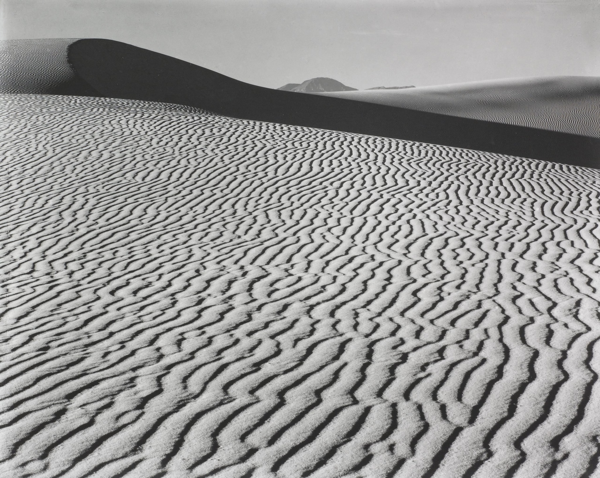 Black and white photo of sand dunes with rippled patterns in the foreground, a dark curved shadow dividing the image, and smooth hills in the distance under a clear sky.