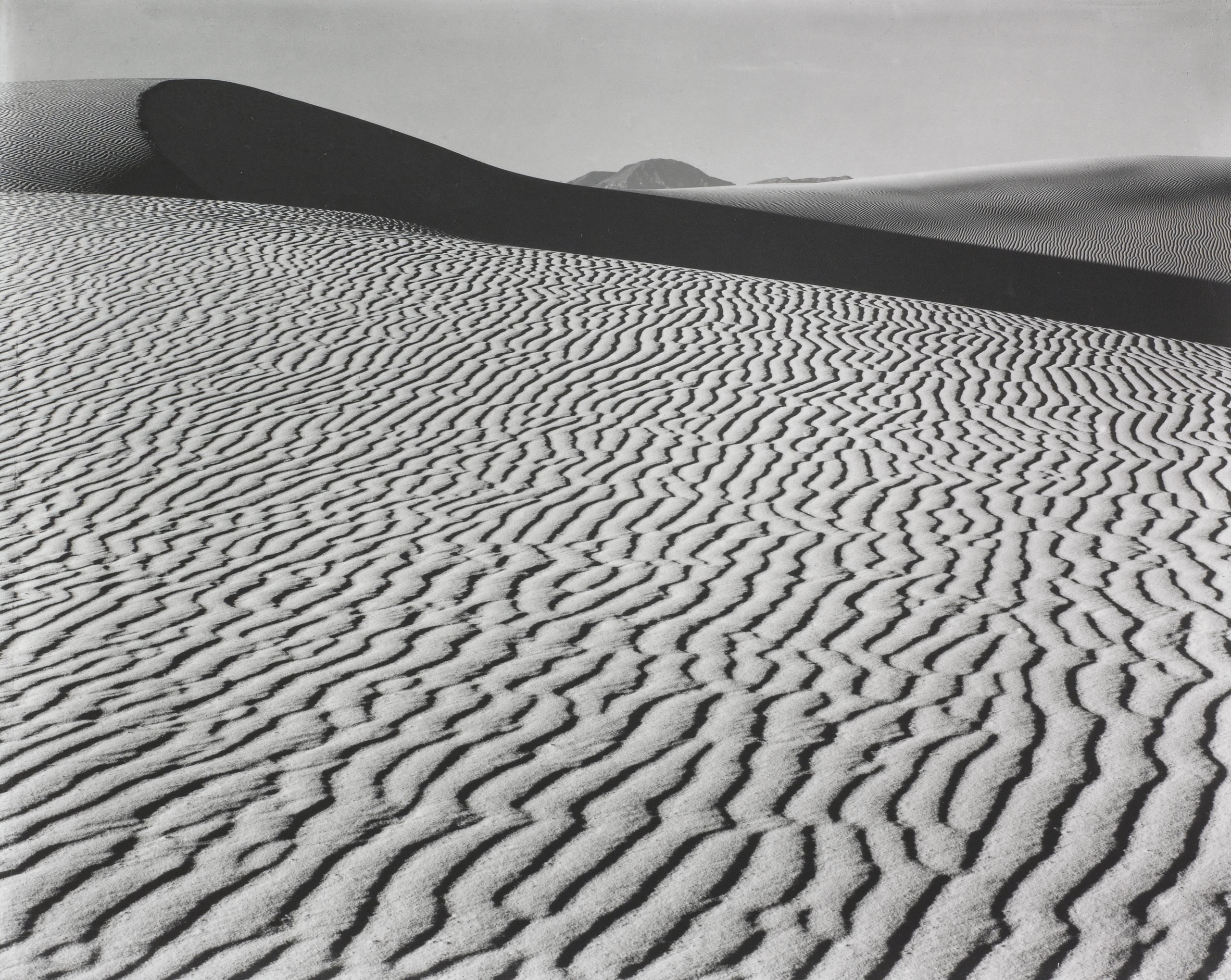 Black and white photo of sand dunes with rippled patterns in the foreground, a dark curved shadow dividing the image, and smooth hills in the distance under a clear sky.