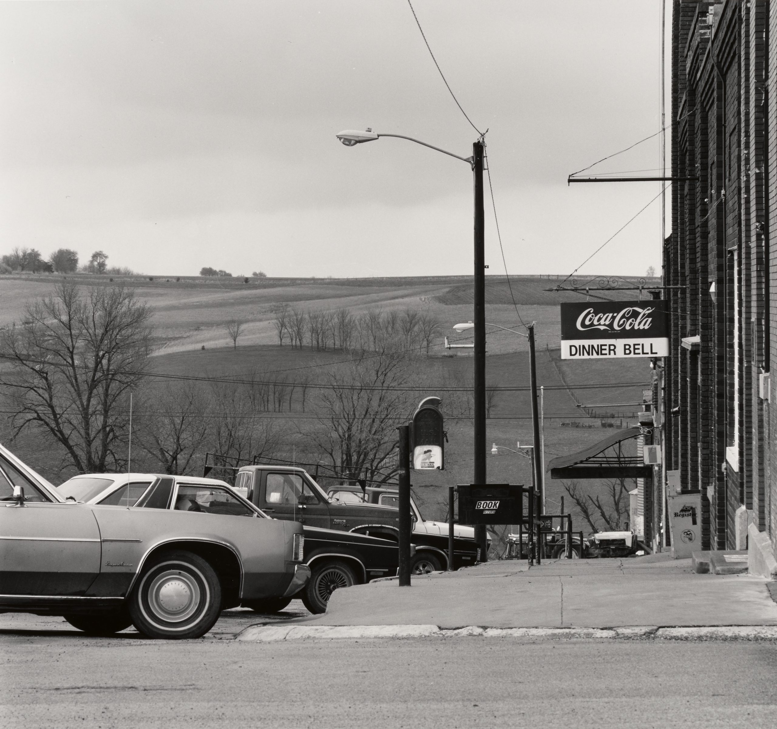 Black and white photo of parked cars on a small-town street beside a sidewalk. A sign reads "Coca-Cola Dinner Bell." Leafless trees and rolling fields are visible in the background under a cloudy sky.