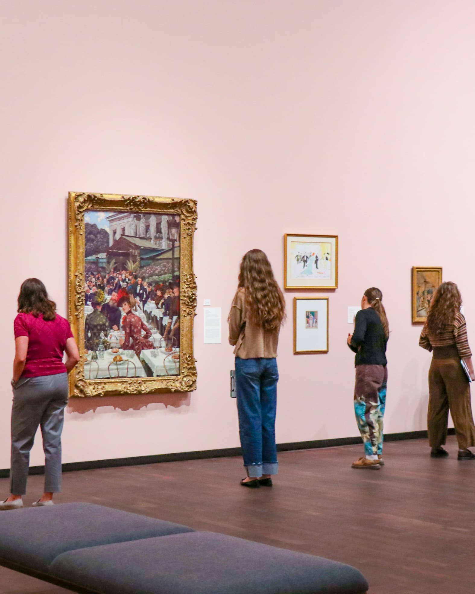 Four people stand in a gallery, observing various paintings on a pink wall. The largest painting has an ornate gold frame; others are smaller with simple frames. The floor is dark wood, and a gray bench is in the foreground.