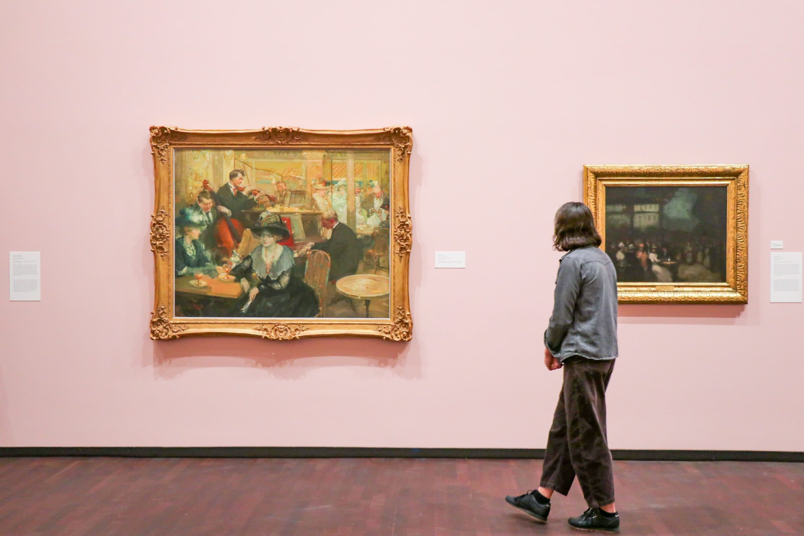 A person with shoulder-length hair views framed paintings on a pink wall in an art gallery. The left painting shows a busy café scene, while the right depicts a darker interior scene.