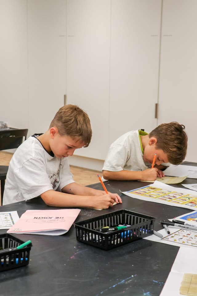 Two young boys sit at a table, focused on drawing or coloring with pencils. Art supplies and papers are spread out in front of them in a classroom setting.
